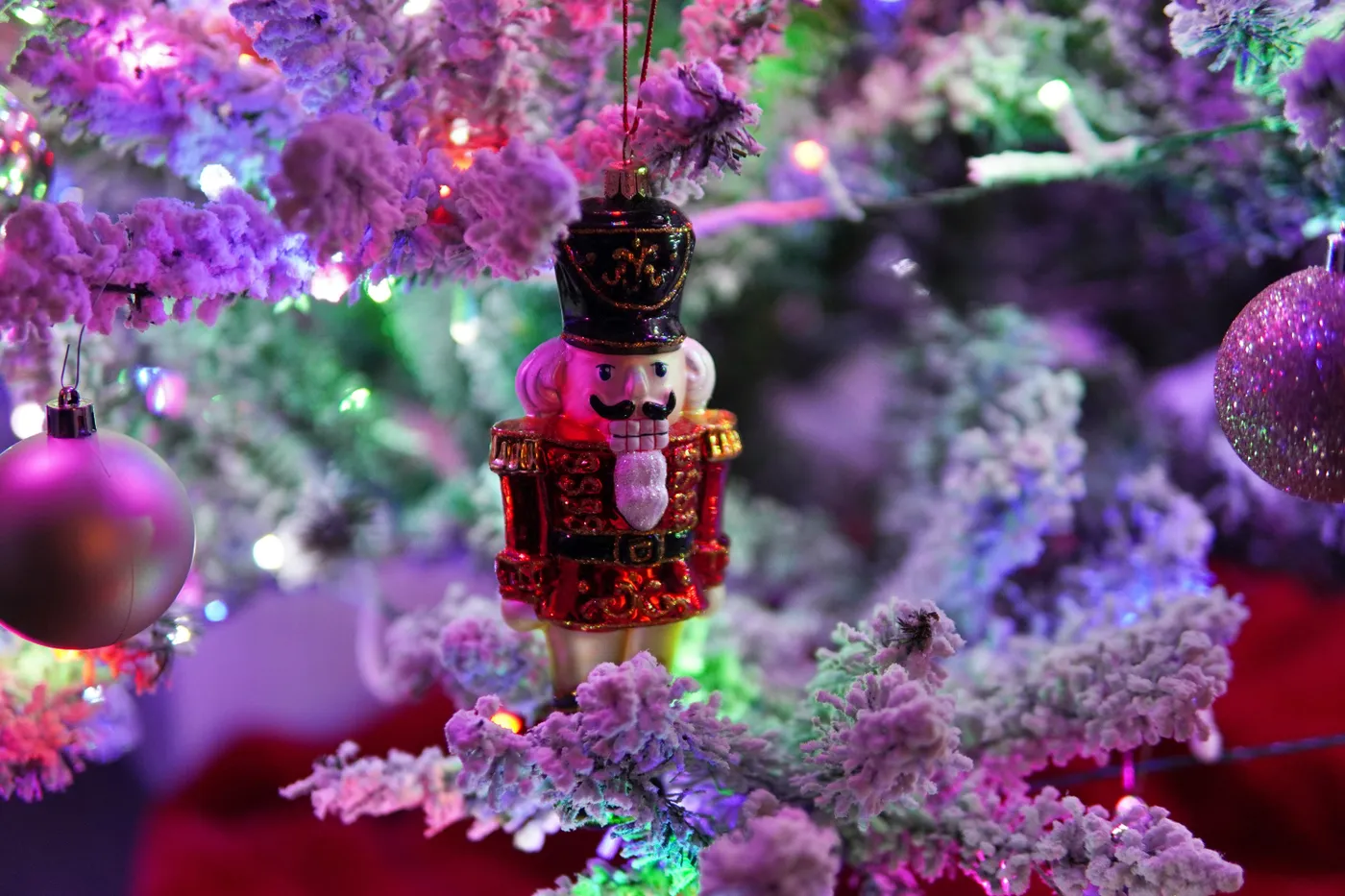 A festive close-up of a nutcracker ornament hanging on a snow-frosted Christmas tree, illuminated by twinkling multicolored holiday lights. The nutcracker, with its intricate red and gold detailing, s