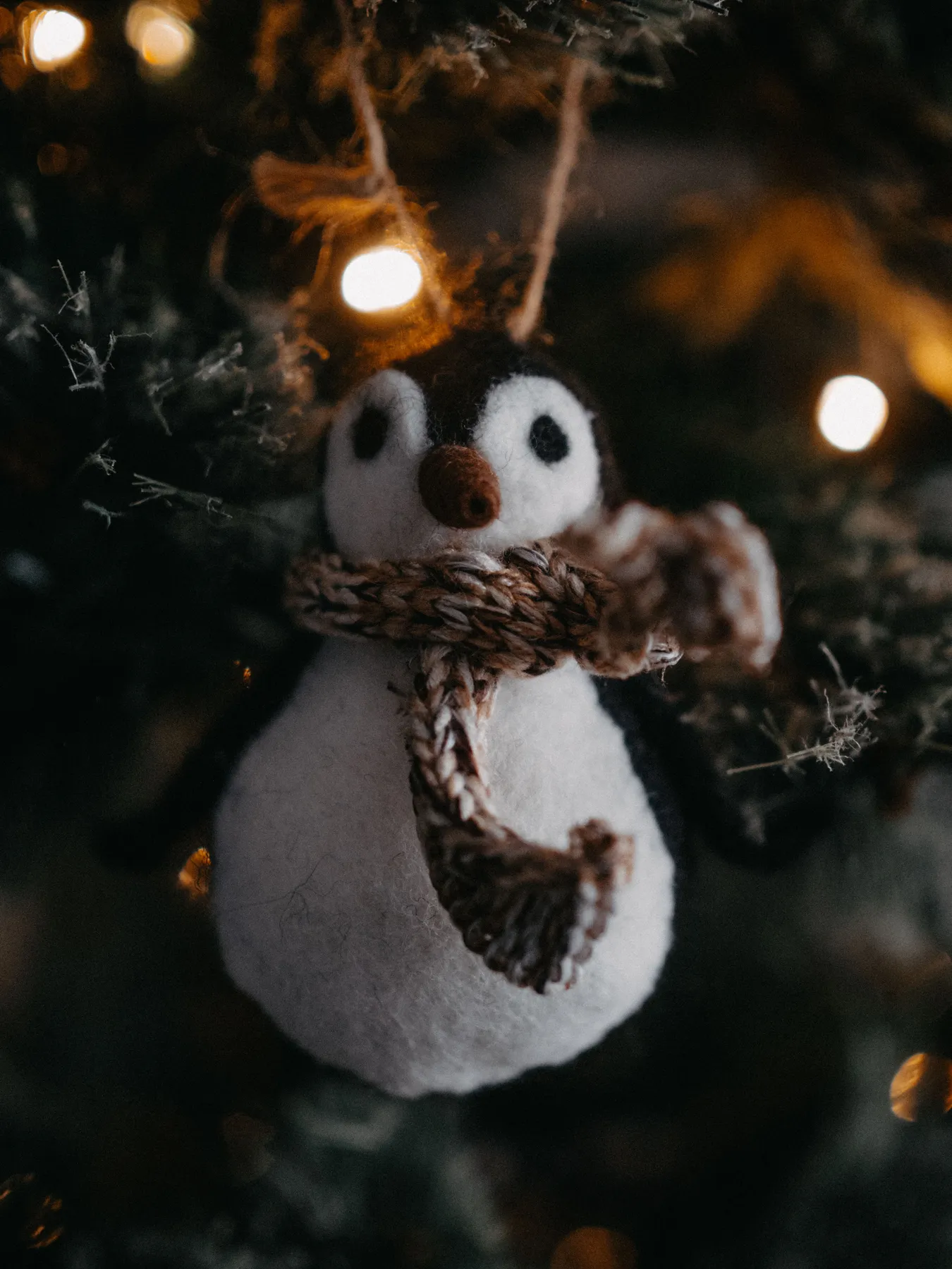 A handmade felt penguin ornament hangs on a Christmas tree with warm festive lights.
