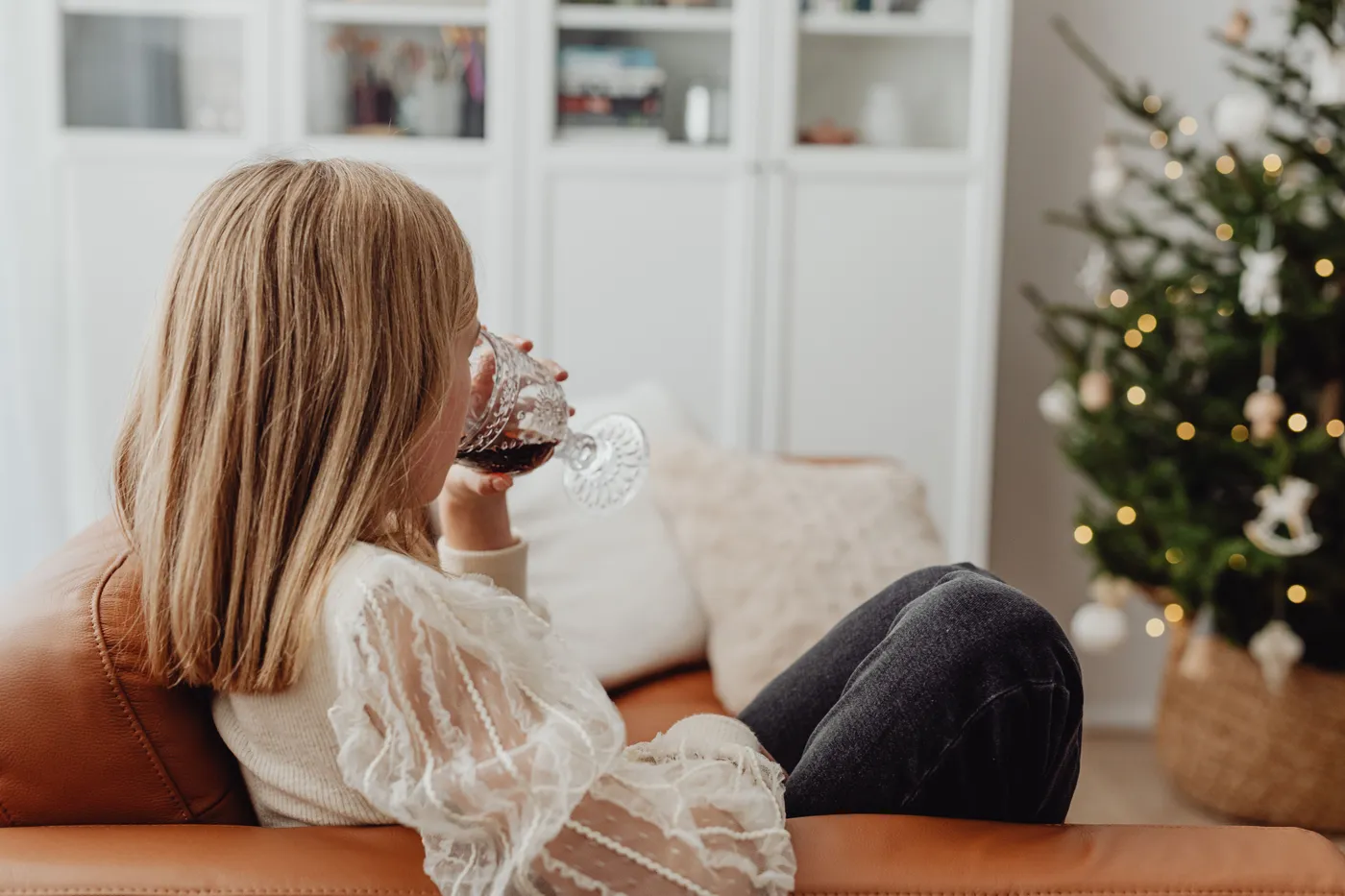 A woman enjoys a glass of wine while sitting by a decorated Christmas tree in a cozy living room.
