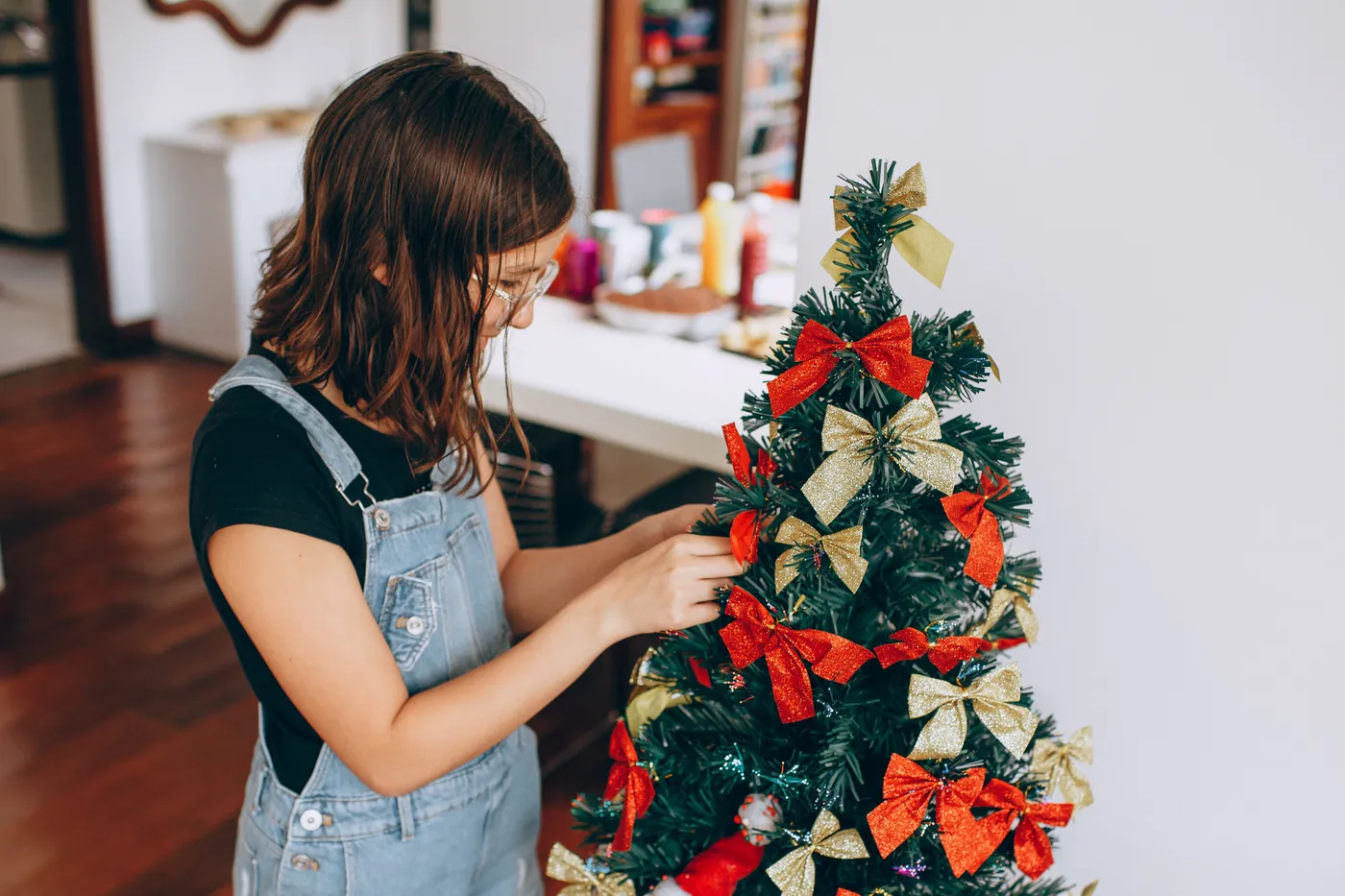 A young girl in overalls decorates a small Christmas tree indoors with festive bows.