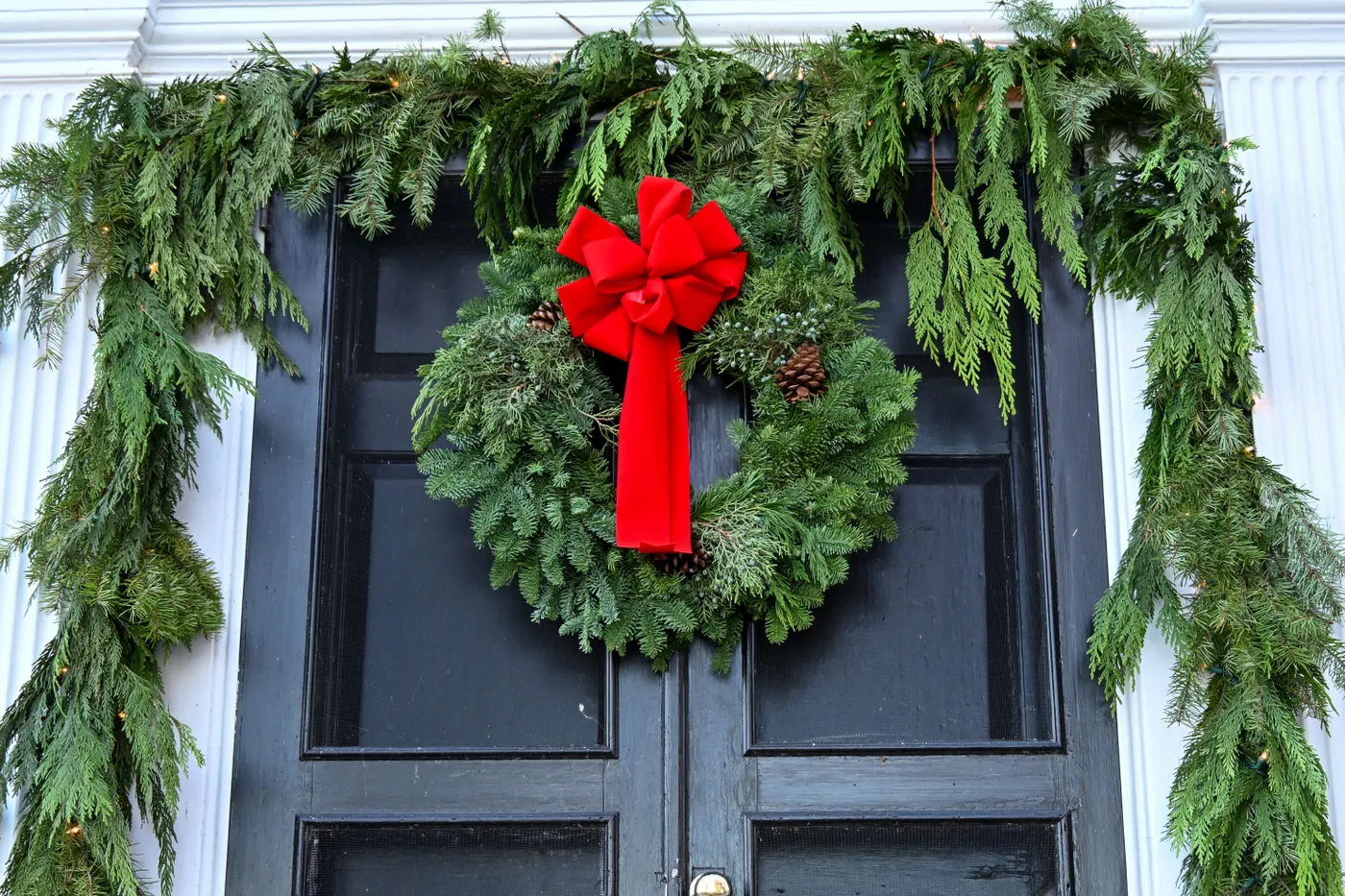 Elegant evergreen wreath with red bow adorns a classic black door in winter season.