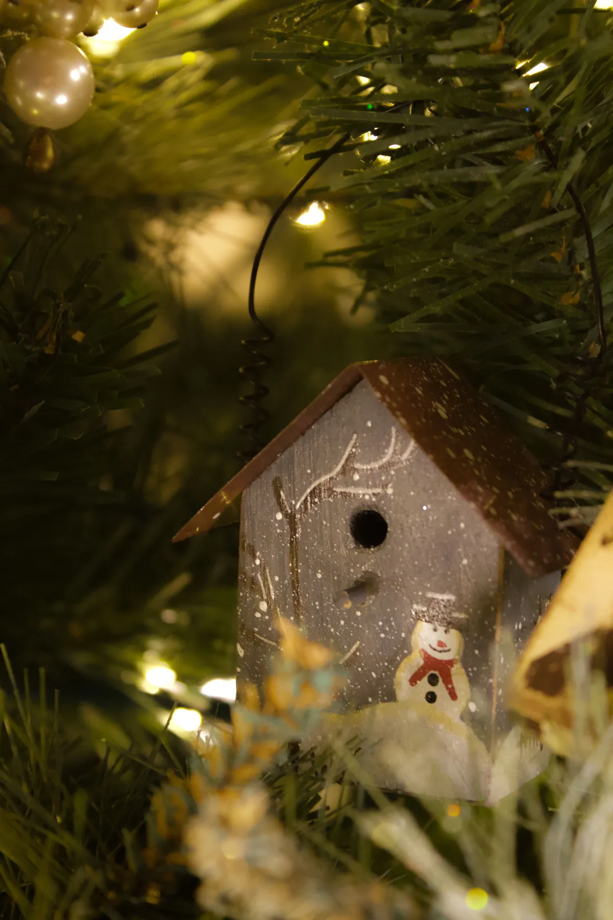 Festive birdhouse ornament nestled among Christmas tree branches and glowing lights.