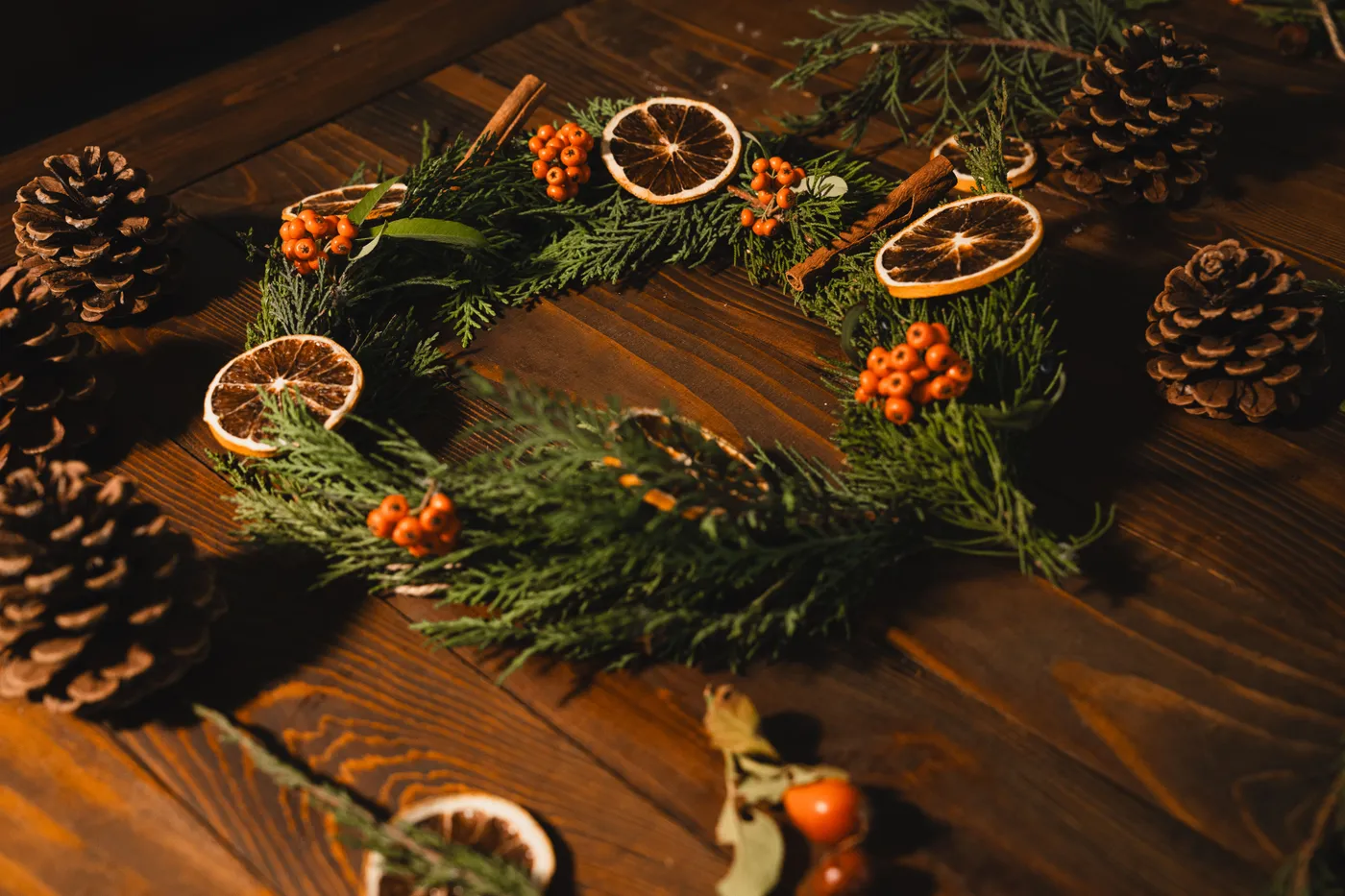Festive holiday wreath made of evergreen, dried citrus, and pinecones on a wooden table.