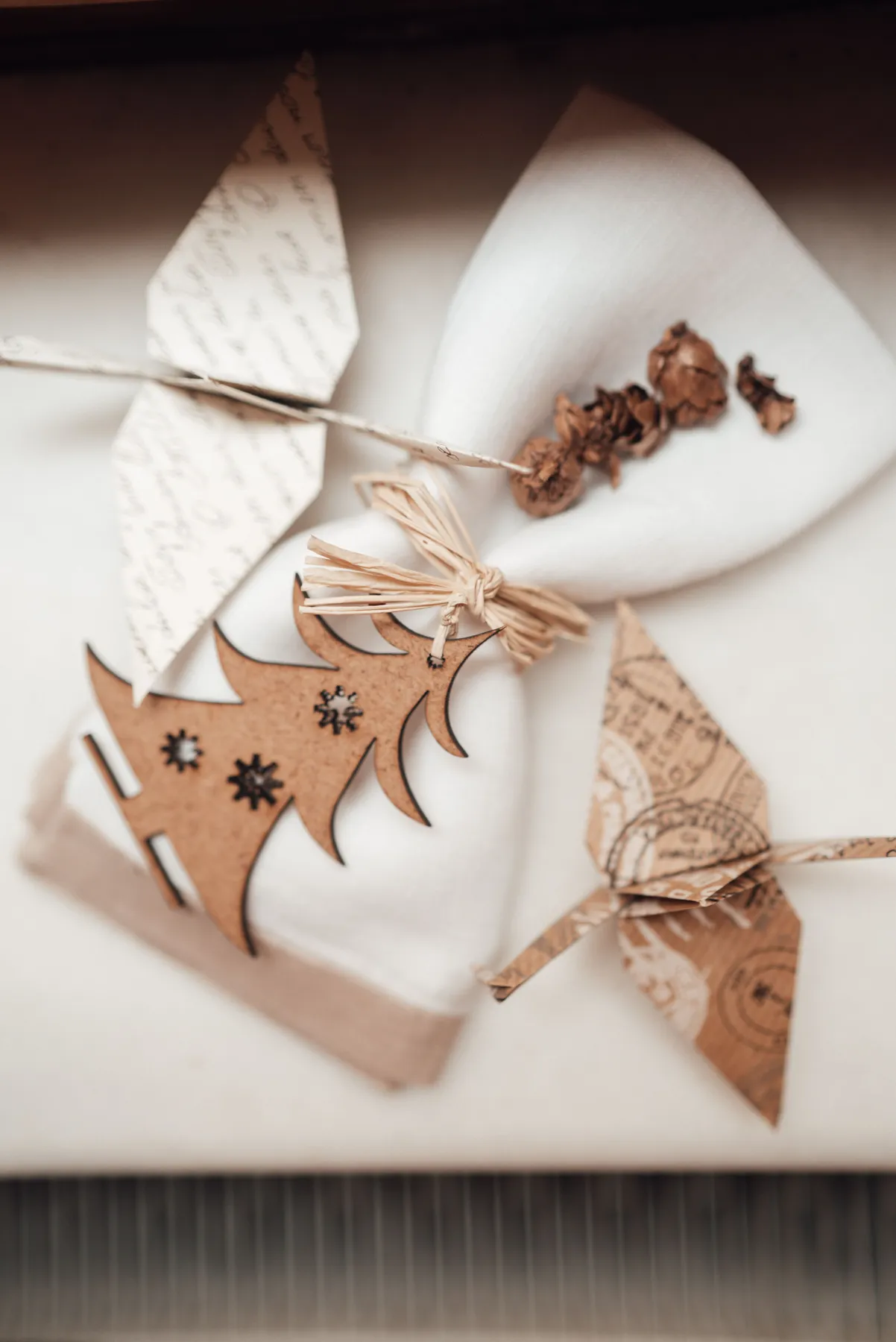 From above of white cloth with napkin covered in rope with small wooden Christmas tree near decorative objects and origami cranes from paper in light place