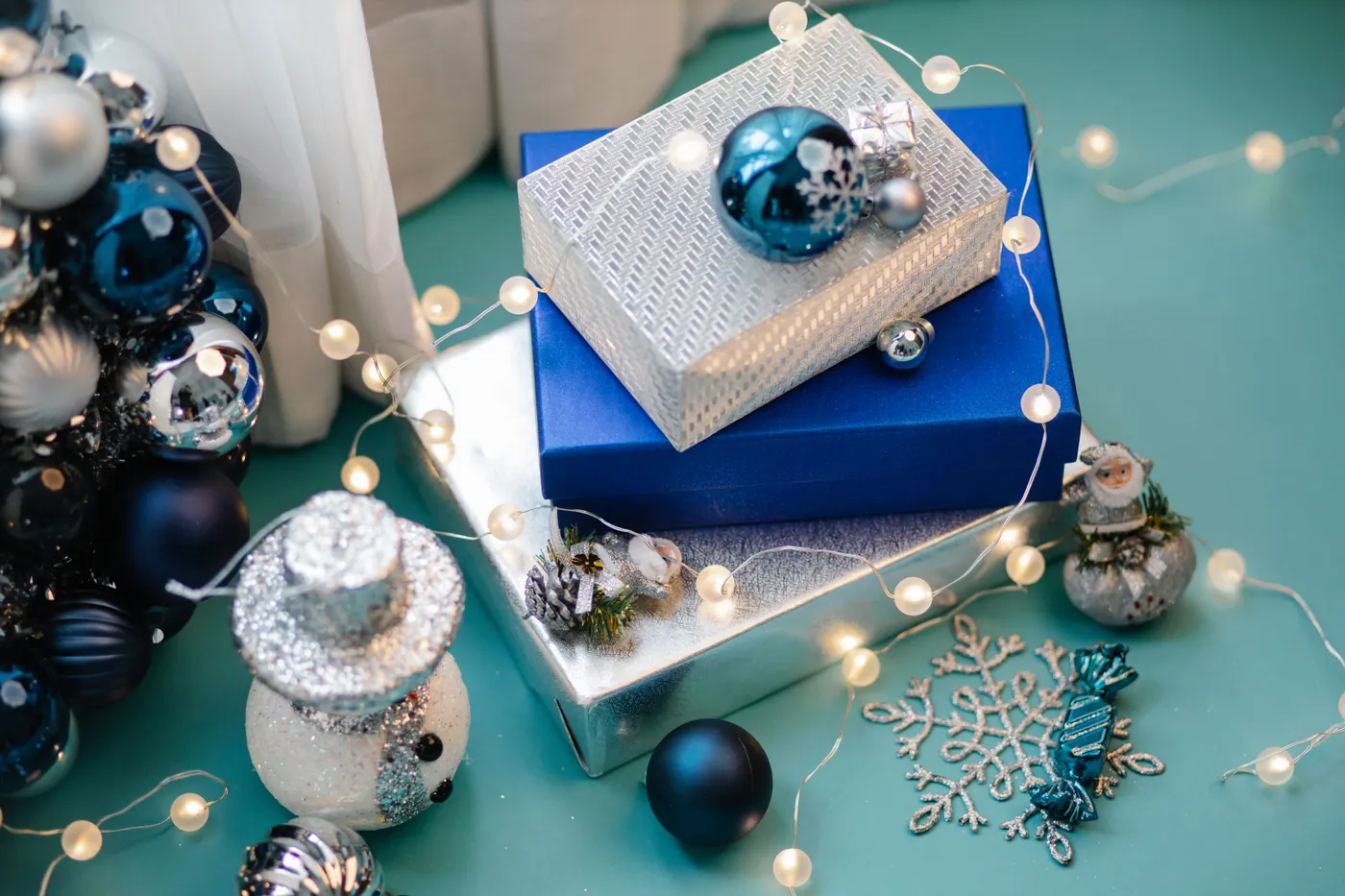 High angle of glowing fairy lights with pile of Christmas gift boxes and toys placed on blue surface of table