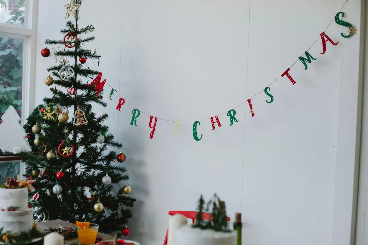 Light room with Christmas tree with balls and toys near window and white wall with words merry Christmas near dinner table with cake and glass with orange drink near chair
