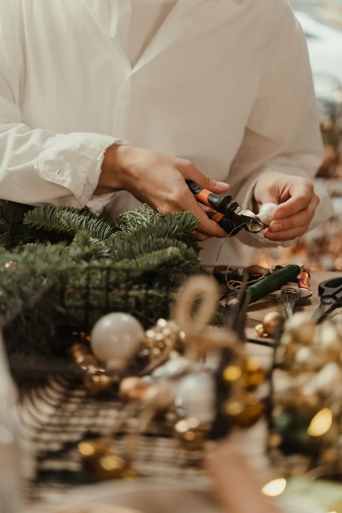 Person crafting a Christmas wreath using pliers and decorations on a festive table.