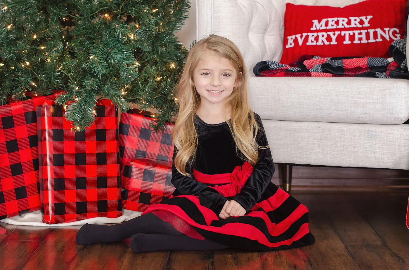 Young girl in black and red dress sitting by a Christmas tree with gifts, spreading holiday cheer.